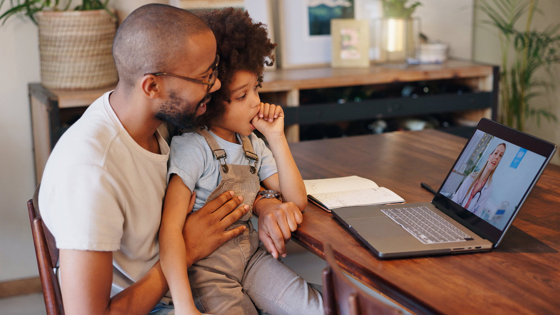 A father holds his young son in his lap while they both look at a laptop with a doctor on the screen for a telehealth visit. The little boy has big curly hair and is sucking his thumb