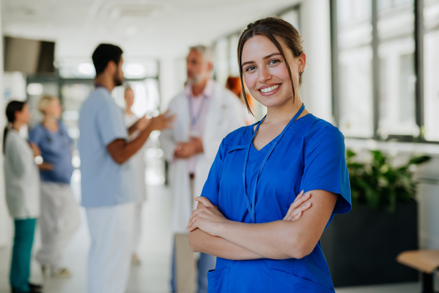 A young female nurse in blue scrubs smiles in a hospital corridor. The blurred out background has a group of nurses and doctors.